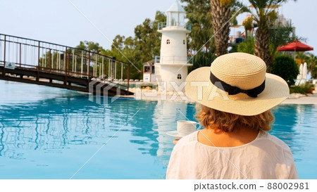 female 50 years old having breakfast by the pool in a straw hat wearing a white dress. woman sitting female 50 years old having breakfast by the pool in a straw hat wearing a white dress. woman sitting 88002981
