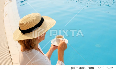 female 50 years old having breakfast by the pool in a straw hat wearing a white dress. woman sitting female 50 years old having breakfast by the pool in a straw hat wearing a white dress. woman sitting 88002986