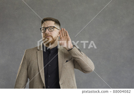 Curious serious man with a worried expression holds his hand to his ear overhearing a secret conversation. Man in suit spies and eavesdrops standing on a gray background. Secret and privacy concept. Curious serious man with a worried expression holds his hand to his ear overhearing a secret conversation. Man in suit spies and eavesdrops standing on a gray background. Secret and privacy concept. 88004082