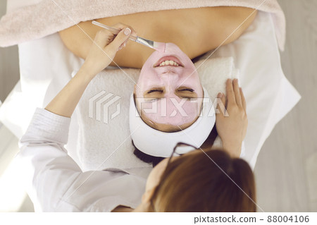 Cosmetology and medical services office. Top view of unknown female beautician with brush applies pink mask on face of satisfied client. Concept of cosmetic procedures in a medical center. 88004106