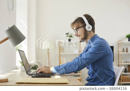 Young happy man in headphones typing on laptop and using internet web online services. Man relaxing and playing games or working remotely at home using modern technology and communication devices. 88004116