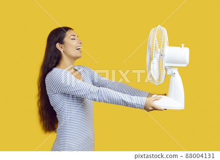 Device for cooling air. Satisfied woman suffering from heat enjoys cold air from electric fan on yellow background. Beautiful smiling woman holding fan in front of her. Hot weather concept. Device for cooling air. Satisfied woman suffering from heat enjoys cold air from electric fan on yellow background. Beautiful smiling woman holding fan in front of her. Hot weather concept. 88004131