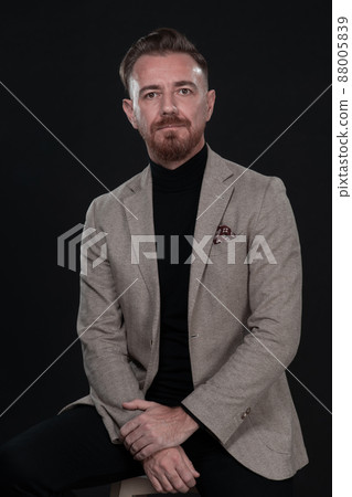 Portrait of adult businessman wearing trendy suit and sitting in modern studio on stylish chair against the black background 88005839