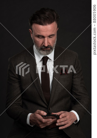 Portrait of adult businessman wearing trendy suit and sitting in modern studio on stylish chair against the black background Portrait of adult businessman wearing trendy suit and sitting in modern studio on stylish chair against the black background 88005906