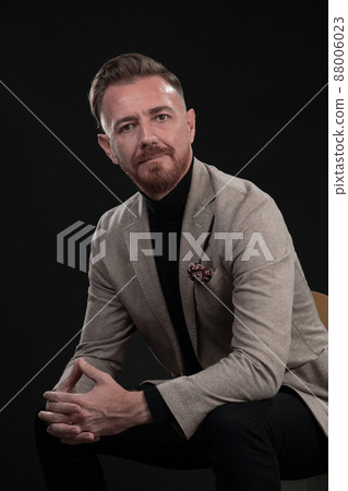 Portrait of adult businessman wearing trendy suit and sitting in modern studio on stylish chair against the black background 88006023