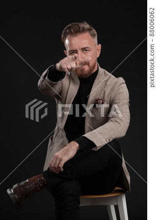 Portrait of adult businessman wearing trendy suit and sitting in modern studio on stylish chair against the black background Portrait of adult businessman wearing trendy suit and sitting in modern studio on stylish chair against the black background 88006062