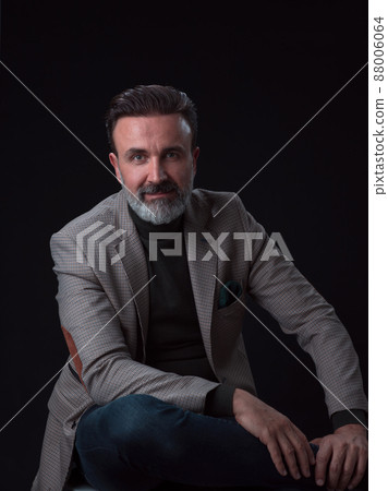 Portrait of adult businessman wearing trendy suit and sitting in modern studio on stylish chair against the black background 88006064