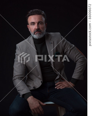 Portrait of adult businessman wearing trendy suit and sitting in modern studio on stylish chair against the black background Portrait of adult businessman wearing trendy suit and sitting in modern studio on stylish chair against the black background 88006072