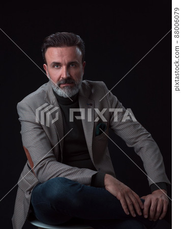 Portrait of adult businessman wearing trendy suit and sitting in modern studio on stylish chair against the black background 88006079