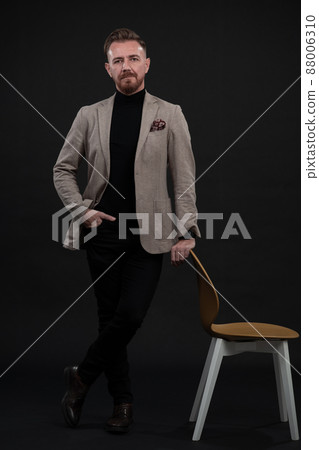 Portrait of adult businessman wearing trendy suit and sitting in modern studio on stylish chair against the black background Portrait of adult businessman wearing trendy suit and sitting in modern studio on stylish chair against the black background 88006310