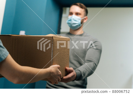 Delivery man wearing medical face mask giving cardboard box parcel to female customer on doorstep at home. Rear view of unrecognizable woman client receiving parcel from courier male at front door 88006752