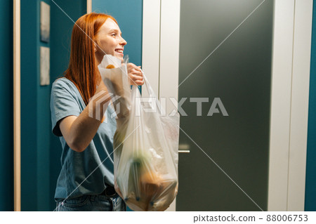 Portrait of cheerful young woman housewife holding groceries bag and smiling looking away, standing on apartment doorstep. Female customer received online order from delivery service. 88006753