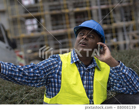 Indian or Somali engineer with safety jacket and helmet talking on mobile phone on construction site 88008139