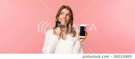 Finance, shopping and technology concept. Close-up portrait of thoughtful, dreamy attractive blond girl in white dress, thinking what buy, order something online shop, hold smartphone and credit card 88010909