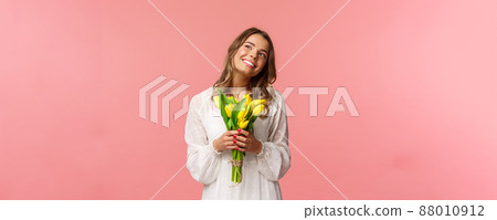 Holidays, beauty and spring concept. Portrait of dreamy, happy blond girl feeling romantic daydreaming about her girlfriend, holding yellow tulips, wear white dress, standing pink background 88010912