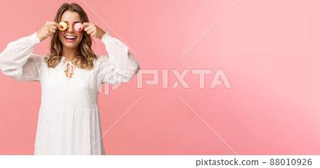 Holidays, spring and party concept. Portrait of carefree funny and cute blond girl in white dress, holding two macarons on eyes and smiling happy, like eating tasty desserts, pink background Holidays, spring and party concept. Portrait of carefree funny and cute blond girl in white dress, holding two macarons on eyes and smiling happy, like eating tasty desserts, pink background 88010926