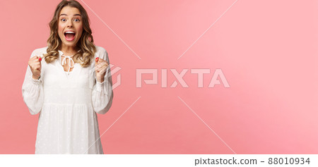 Portrait of surprised lucky hopeful, blond girl winning, hear great news and chanting, gasping open mouth smiling joyful, jumping from happiness and joy, standing pink background Portrait of surprised lucky hopeful, blond girl winning, hear great news and chanting, gasping open mouth smiling joyful, jumping from happiness and joy, standing pink background 88010934