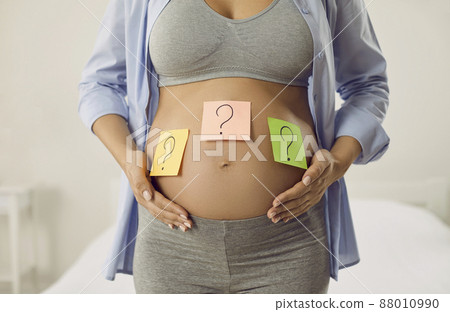 Happy woman thinking of a name or trying to guess the gender of her unborn baby. Cropped shot of a pregnant lady with question mark post it notes on her big belly. Name choice, gender reveal concept Happy woman thinking of a name or trying to guess the gender of her unborn baby. Cropped shot of a pregnant lady with question mark post it notes on her big belly. Name choice, gender reveal concept 88010990