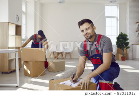 Moving service workers pack cardboard boxes and checking list in client's apartment. Portrait of smiling man in overalls who writes something in clipboard. Delivery and moving day concept. 88011012