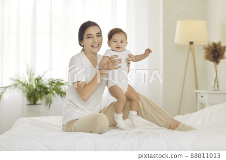 Portrait of smiling mother sit on bed at home play with cute small newborn baby together. Happy caring mom and little infant kid show love and enjoyment. Motherhood and childcare concept. 88011013