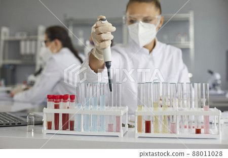 Scientist working in modern pharma or biotech laboratory. Serious woman in PPE mask and goggles doing research and using pipette to transfer liquid in glass tubes. Soft focus close up, science concept 88011028