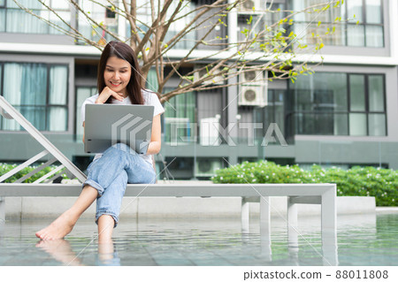Happy young entrepreneur woman sitting on tanning bed beside pool and using laptop computer for remote online working digital, online business project in quiet yard of resort house, Work on vacation 88011808