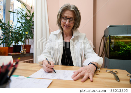 Stylish elegant inspired elderly female fashion designer dressmaker tailor holding a pencil and drawing sketches for clothes, developing new line of dress collection in her design studio Stylish elegant inspired elderly female fashion designer dressmaker tailor holding a pencil and drawing sketches for clothes, developing new line of dress collection in her design studio 88013895