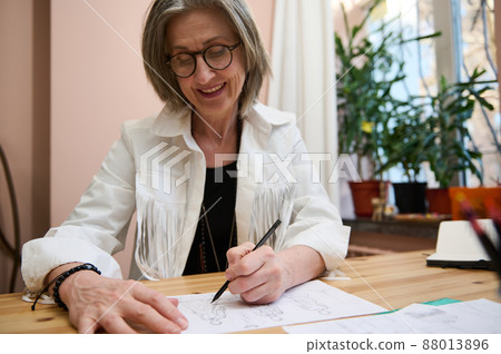 Fashion designer tailor dressmaker, elderly business woman drawing sketches for clothes, creating a new line of dress collection in her cozy creative fashion design studio 88013896