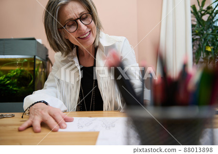 View through blurred colorful pencils holder to a fashion designer, dressmaker, mature elegant Caucasian business woman working at wooden desk, drawing sketches for clothes in her creative workshop 88013898