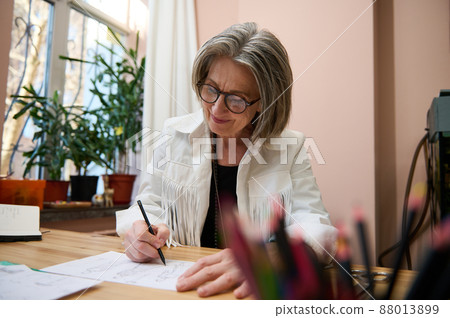 View through blurred pencils holder to a female fashion designer, dressmaker, senior elegant Caucasian business woman working at wooden desk, drawing sketches for clothes in her creative workshop View through blurred pencils holder to a female fashion designer, dressmaker, senior elegant Caucasian business woman working at wooden desk, drawing sketches for clothes in her creative workshop 88013899