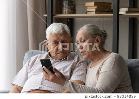 Senior couple discuss mobile app seated on sofa at home Senior couple discuss mobile app seated on sofa at home 88014859
