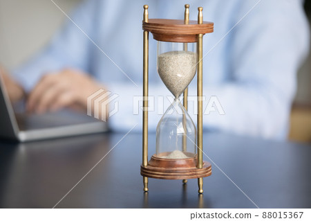 Closeup hourglass on table, businesswoman working on laptop on background Closeup hourglass on table, businesswoman working on laptop on background 88015367