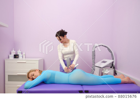 A female client receives a LPG massage in a beauty clinic. Beauty parlour. Blue suit. Lilac background. 88015676