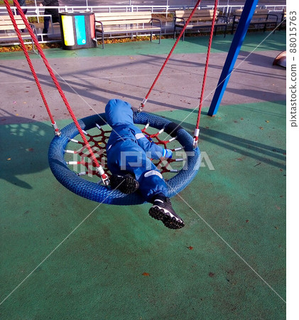 A little boy in a blue jumpsuit swinging on a swing on the playground in spring or autumn. A little boy in a blue jumpsuit swinging on a swing on the playground in spring or autumn. 88015763