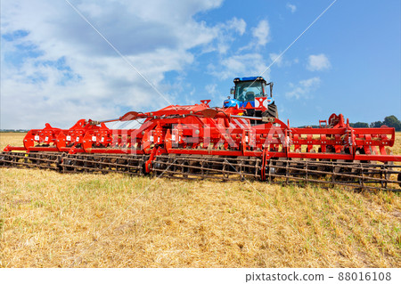 Universal red harrow as a hitch on an agricultural tractor against the background of a compressed yellow wheat field. 88016108