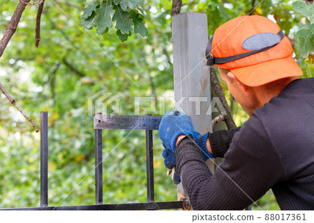 The worker sets a right angle on the fence with a metal corner. The worker sets a right angle on the fence with a metal corner. 88017361