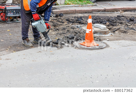 A worker with an electric jackhammer breaks the old asphalt from the road around the sewer manhole. A worker with an electric jackhammer breaks the old asphalt from the road around the sewer manhole. 88017362