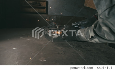 Forge workshop. Smithy. Worker in a welding hood helmet welds a part by electric welding. Sparks are reflected in the protective screen. Forge workshop. Smithy. Worker in a welding hood helmet welds a part by electric welding. Sparks are reflected in the protective screen. 88018191