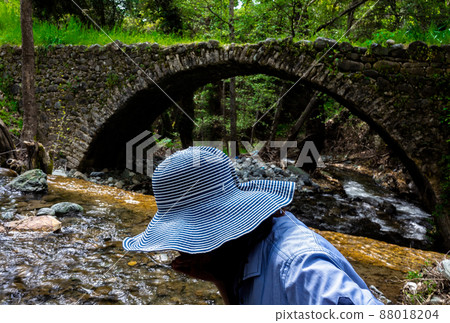 A girl in a wide-brimmed hat drinks water from a stream next to a medieval Venetian bridge in the center of the island of Cyprus. A girl in a wide-brimmed hat drinks water from a stream next to a medieval Venetian bridge in the center of the island of Cyprus. 88018204