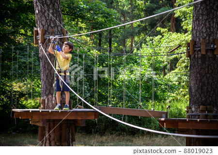 Strong excited young boy playing outdoors in rope park. Caucasian child dressed in casual clothes and sneakers at warm sunny day. Active leisure time with children concept Strong excited young boy playing outdoors in rope park. Caucasian child dressed in casual clothes and sneakers at warm sunny day. Active leisure time with children concept 88019023