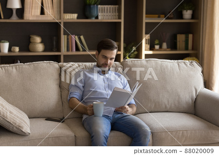 Happy young guy read good book drinking tea on sofa Happy young guy read good book drinking tea on sofa 88020009