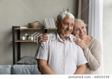 Elderly loving couple standing in living room looking out window 88020199