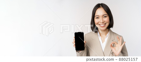 Smiling asian woman showing smartphone screen and okay sign. Corporate person demonstrates mobile phone app interface, standing over white background Smiling asian woman showing smartphone screen and okay sign. Corporate person demonstrates mobile phone app interface, standing over white background 88025157