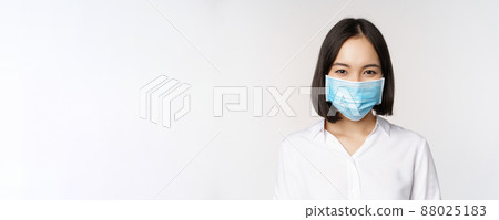 Covid and healthcare concept. Close up portrait of asian woman, office lady in face mask, smiling, using protection from coronavirus during pandemic, white background 88025183
