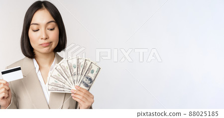 Thoughtful businesswoman, korean corporate woman showing credit card and money cash, dollars in hands, standing over white background and thinking 88025185