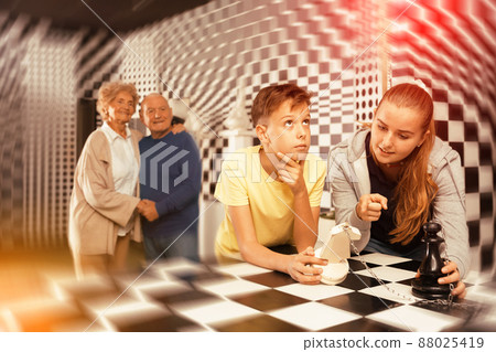 Kids solving chess riddle in escape room and grandparents watching them Kids solving chess riddle in escape room and grandparents watching them 88025419