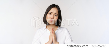 Close up portrait of asian woman begging, asking for smth, say please with coy sad face, need help, standing over white background 88025788