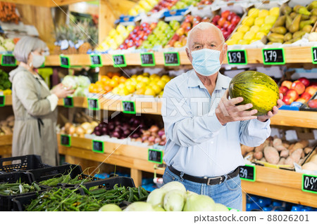 Senior man in mask choosing melon in greengrocer Senior man in mask choosing melon in greengrocer 88026201