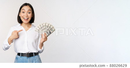 Portrait of young asian woman pointing at her money dollars, showing cash, standing over white background Portrait of young asian woman pointing at her money dollars, showing cash, standing over white background 88026629