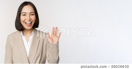 Friendly business woman, asian office lady waving hand and saying hello, hi gesture, standing over white background Friendly business woman, asian office lady waving hand and saying hello, hi gesture, standing over white background 88026630